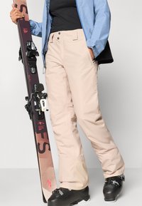 Μπεζ σκι παντελόνι με ίσια κόψιμο, με δύο μπροστινές τσέπες και αντίθετης μαύρης σκελετικής. Φορεμένο με μπότες σκι και κρατώντας σκι.