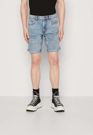 Shorts in denim con un lavaggio blu chiaro, con risvolti, design a cinque tasche, lievi segni di usura, abbinati a sneakers nere alte e calze.
