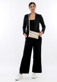 Borsa a tracolla beige con tasca anteriore con zip e nappina, indossata su un outfit nero e abbinata a sneaker bianche. Design semplice e funzionale.