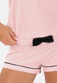 Růžové pyžamo s bílými puntíky, černými akcenty a mašlí v pase; lehký materiál s volným střihem a krátkými rukávy.