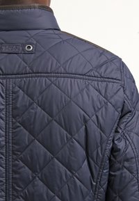 Marineblå quiltet jakke med høj krave og synlige sømme. Indeholder en mærkedetalje nær kraven og tekstureret stof.