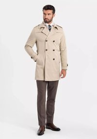 Beżowy dwurzędowy płaszcz trench z czarnymi guzikami, z wykładanym kołnierzem i bocznymi kieszeniami, w połączeniu z ciemnobrązowymi spodniami i butami.