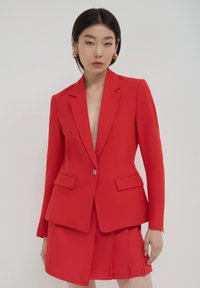 Femme portant un blazer rouge ajusté avec un bouton unique et une jupe plissée assortie, accessoirisée avec des boucles d'oreilles en or.