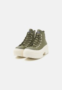 Botas de plataforma altas de color verde aceituna con suelas gruesas blancas, cordones y suela resistente, aisladas sobre un fondo blanco.