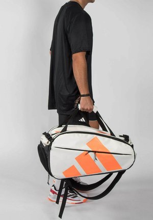 Mann in schwarzem Hemd und Shorts hält eine große weiße und orangefarbene Adidas-Sporttasche an den Griffen vor einfarbigem Hintergrund.