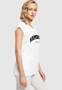 Hvit T-skjorte med svart "HAMBURG" tekst, kort erm, og V-hals design. Brukt med lysfargede bukser. Glatt stofftekstur.
