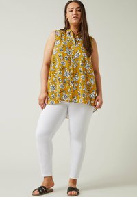 Blusa senza maniche gialla a fiori con motivi neri, con bottoni frontali e orlo asimmetrico, abbinata a jeans skinny bianchi e sandali neri.