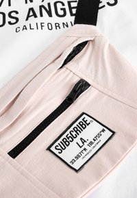 Lyserød skuldertaske med lynlåsrum, sorte beslag og et mærke med teksten "SUB5CRIBE" under et hvidt og sort grafisk design.