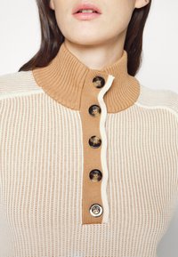 Zbliżenie na kobietę w kremowym i brązowym swetrze ściąganym, z brązowym kołnierzykiem zapinanym na guziki i pięcioma guzikami w kolorze skorupy żółwia na przodzie.