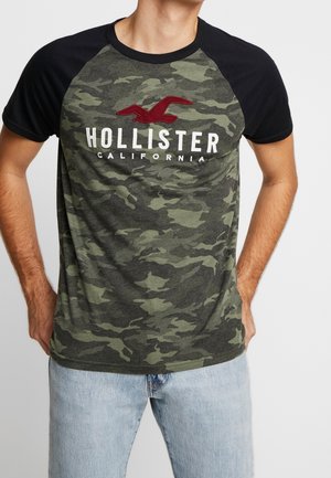 Tričko s krátkým rukávem s maskáčovým vzorem v zelené a černé barvě, s černým límcem a červeným vyšívaným logem "Hollister California."