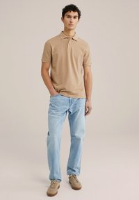 Polo de color marrón claro con cuello y dos botones, combinado con jeans de color azul claro y zapatillas beige. El modelo está de pie con una mano en el bolsillo.