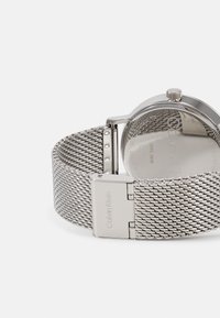 Montre à bracelet en maille argentée avec un boîtier en acier inoxydable rond, design minimaliste et nom de la marque gravé sur le fermoir.
