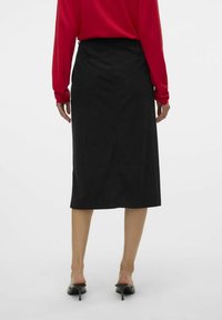 Černá sukně po kolena ve slušivém střihu s jemnou texturou, se zadním rozparkem a páskem. Kombinována s červeným topem s dlouhým rukávem a černými muly na podpatku.