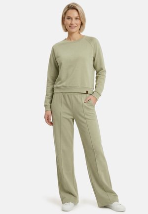 Blondynka stojąca, ubrana w pasującą jasnozieloną bluzę z długim rękawem i spodnie o szerokich nogawkach z białymi trampkami, uśmiechająca się z ręką w kieszeni.