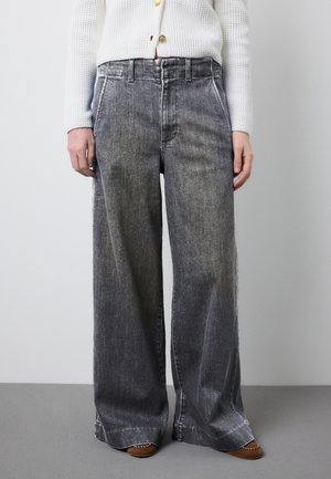 Person trägt weite graue Jeans mit hohem Bund und Seitentaschen, kombiniert mit einem weißen, strukturierten Pullover und braunen Mokassins.