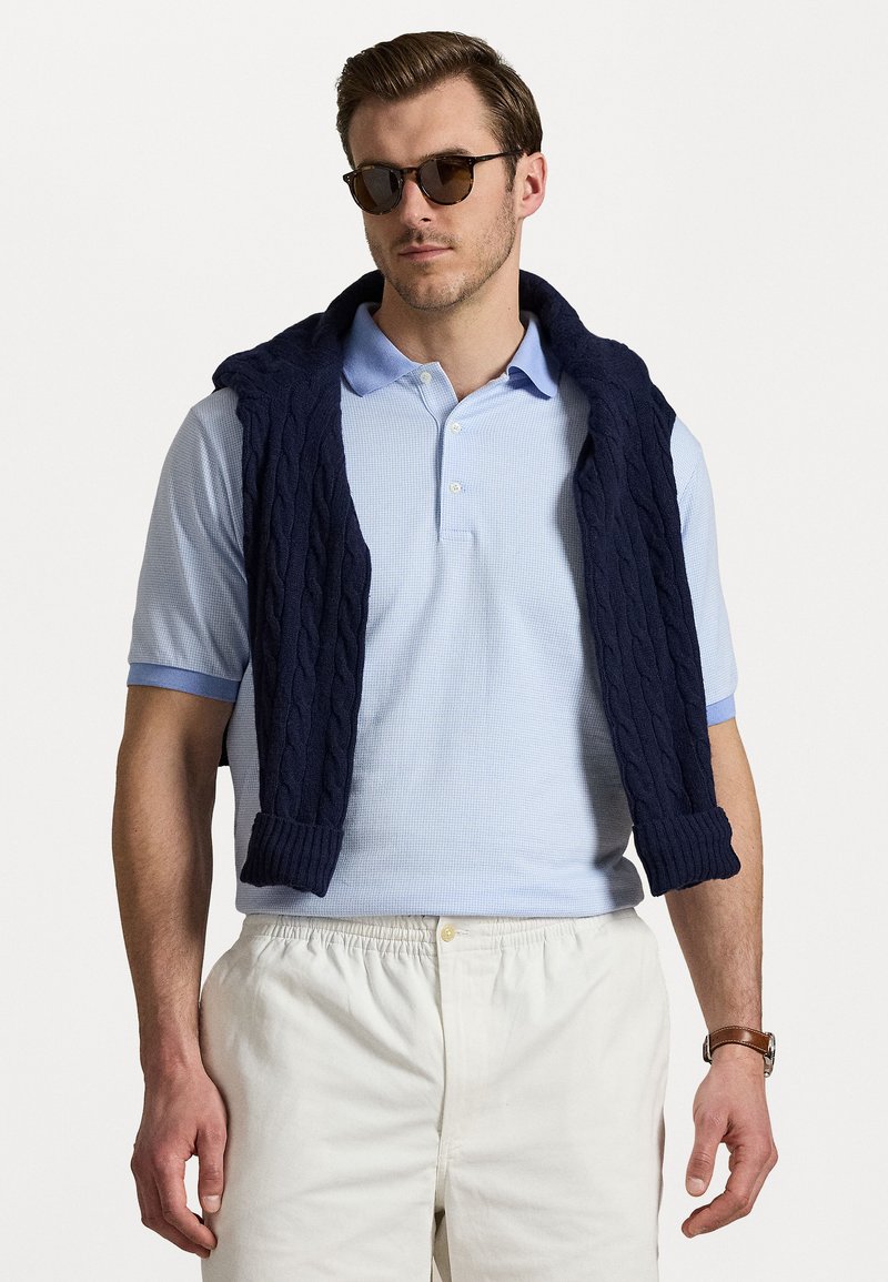 Pulover din tricotaj cu model de împâslire, de culoare bleumarin, așezat peste un tricou polo de culoare bleu deschis. Șorturi de culoare deschisă. Ochelari de soare pe un bărbat cu păr scurt.