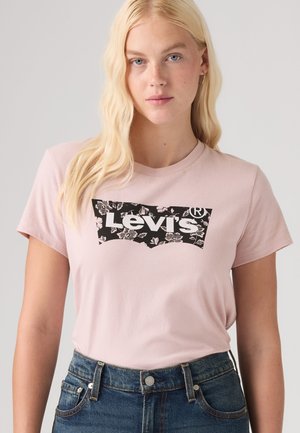 Femme blonde portant un t-shirt Levi's rose pâle avec un logo floral noir, associé à un jean en denim bleu taille haute.