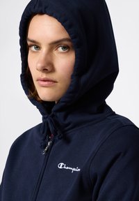 Sudadera con cremallera de color azul marino, fabricada en un tejido suave, que cuenta con una capucha ajustable con cordón y un pequeño logo blanco en el pecho.