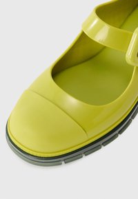 Green, glossy Mary Jane-style shoe with a rounded toe, strap across the foot, and textured rubber sole. Features matte accents at the front.