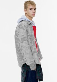 Grå mønstret jakke med denim tekstur, lagdelt over en rød sweatshirt og en grønternet skjorte, med hætte og afslappet pasform.