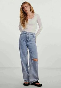 Top de manga longa branca com textura canelada, combinado com jeans de perna larga de cintura alta em azul claro, com rasgos nos joelhos. Modelo usa sandálias pretas.