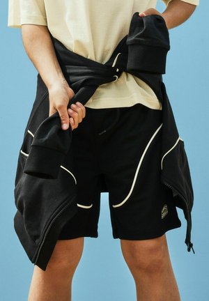 Personne attachant une veste zippée noire autour de la taille, portant une chemise beige à manches courtes et un short noir avec bordure blanche, sur fond bleu.