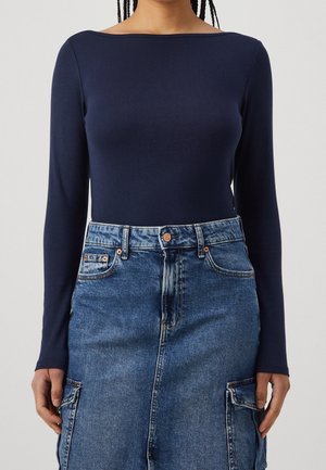 Personne portant un haut bleu marine à manches longues, rentré dans une jupe en denim bleue taille haute avec des poches avant et une fermeture à boutons.