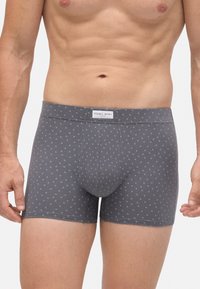 Torso masculino que lleva calzoncillos grises con pequeños patrones de triángulos blancos y una etiqueta de la marca Ysabel Mora en la cintura.