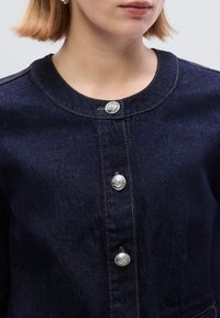 Σκούρο τζην μπουφάν με στρογγυλεμένο γιακά, που διαθέτει ασημ ένια κουμπιά και αντίθετη ραφή. Ομαλή υφή με κλασικό σχέδιο.