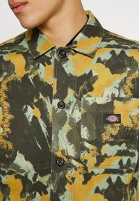 Skjortjacka i olivgrön och senapsgul med mönster, med en bröstficka och svarta knappar. Har en märkeslapp på fickan.