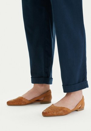 Stopy w beżowych, perforowanych balerinach na płaskim obcasie ze slingbackiem, w połączeniu z ciemnoniebieskimi spodenkami podwiniętymi na jasnym tle.