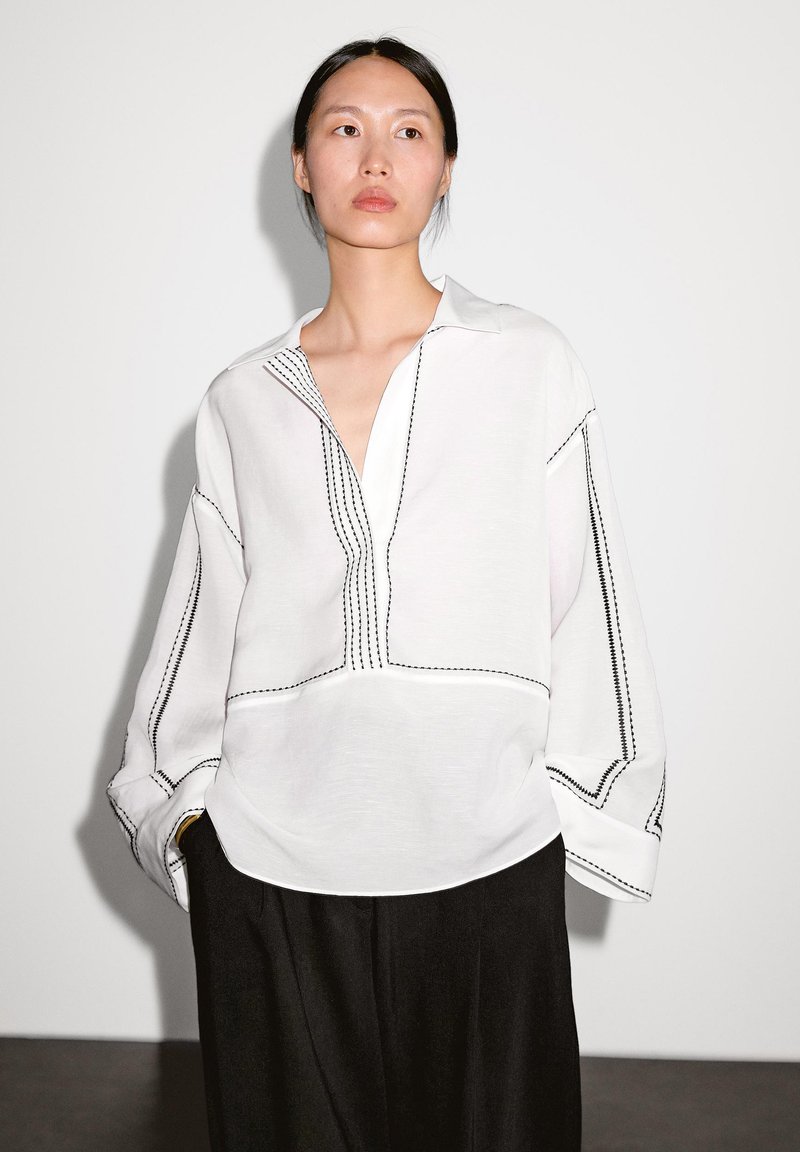 Chemise blanche à manches longues avec col, ornée de coutures noires et d'une coupe décontractée. Texture lisse, avec un décolleté profond et des fentes sur les côtés.