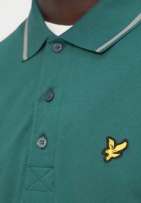 Πράσινο πόλο μπλουζάκι με γιακά που διπλώνει προς τα κάτω, ρίγες σε αντίθετο γκρι χρώμα, τρία σκουρόχρωμα κουμπιά και κεντητό λογότυπο με κίτρινο πουλί στο στήθος.