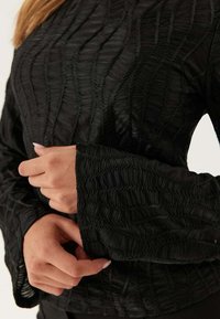 Top nero tessuto con motivi ondulati, maniche lunghe a campana e dettagli intricati. Il materiale sembra leggero e leggermente lucido.