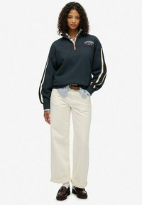 Sweatshirt zippé marine avec des manches rayées, arborant un logo sur la poitrine, associé à un pantalon large crème et des mocassins foncés.