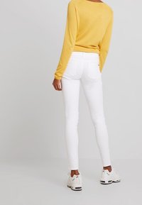 Bílé skinny džíny v kombinaci se žlutým svetrem s dlouhým rukávem, s lodičkovým výstřihem a ribbovanou strukturou, doplněné bílými teniskami.