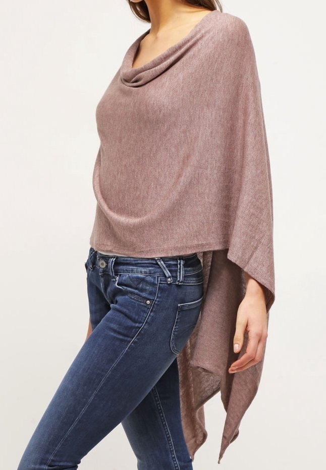 Femme portant un poncho asymétrique mauve avec un col drapé et un jean skinny bleu, debout devant un fond neutre.