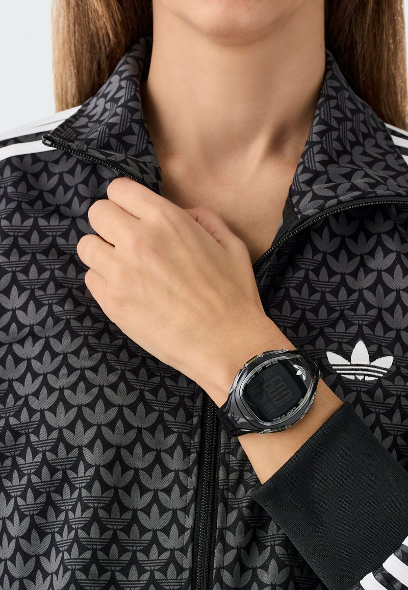 Schwarze Digitaluhr an einem Handgelenk, mit abgerundeter Form und sichtbarem Bildschirm. Getragen mit einer schwarz-grauen Jacke mit Reißverschluss und Muster.