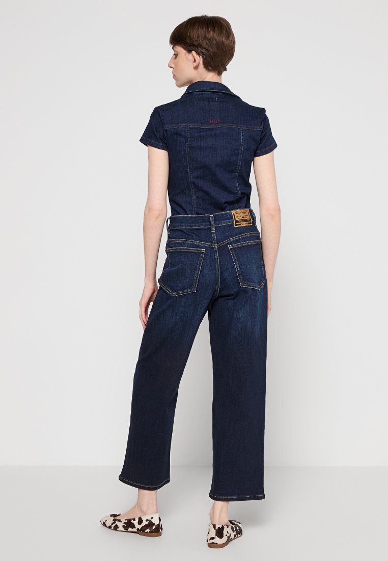 Denim jumpsuit in dark blue with short sleeves, a collar, and back pockets. Features a slightly loose fit and visible stitching accents.