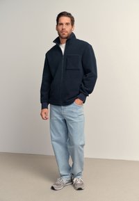 Tamsiai mėlynas džemperis su užtrauktuku, aukštu apykakle ir priekinė kišene, derinamas su šviesiai mėlynais džinsais ir pilkomis sportinėmis avalynėmis neutraliame fone.