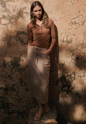 Hosszú hajú nő áll egy repedezett, texturált fal előtt, barna rövid ujjú felsőt, bézs megkötős midi szoknyát és szandált visel, az egyik kezét a zsebében tartja.