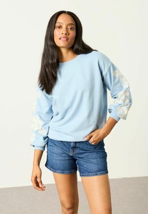 Woman with long dark hair wearing a light blue sweatshirt with white floral embroidery on sleeves and denim shorts, standing with one hand in pocket.