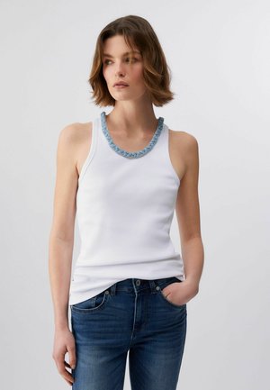 Lühikeste pruunide juustega naine, kes kannab valget varrukateta topi, sinist helmestega kaelakeed ja tumedat sinist teksapükse, seisab ühe käega taskus.