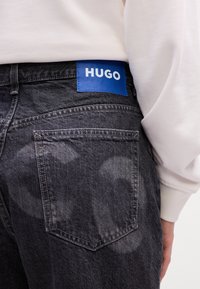 Person med sorte slidte denimjeans, der har en blå "HUGO" etiket på linningen, og en hvid langærmet skjorte.