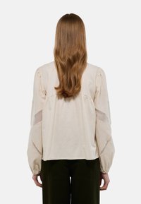 Blouse beige avec des manches bouffantes, des détails en œillets sur les côtés et une coupe décontractée. Fabriquée en tissu léger et texturé.