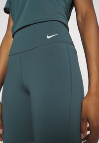 Tmavě petrolejové sportovní šortky s vysokým pasem mají ploché švy a malé bílé logo ve tvaru háčku. Hladká textura, přiléhavý střih.