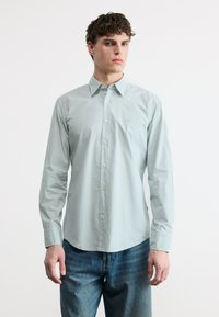 Νέος άνδρας φορώντας ένα γαλάζιο πουκάμισο με κουμπιά και το λογότυπο "BOSS" στο στήθος και ξεθωριασμένο μπλε τζιν, στέκεται μπροστά σε ένα απλό λευκό φόντο.