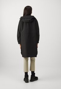 Manteau long noir avec une capuche et des poignets élastiques, présentant un design épuré. Porté sur un pantalon cropped de couleur claire et des chaussures noires.