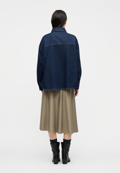 Γυναίκα που στέκεται με την πλάτη, φοράει σκουρό μπλε τζιν μπουφάν, μπεζ midi φούστα, μαύρα μποτάκια αστραγάλου και σκοτεινές κάλτσες σε απλό φόντο.