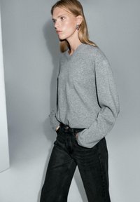 Pullover grigio a collo a V con vestibilità rilassata e maniche lunghe, abbinato a jeans neri con vita alta, su uno sfondo neutro.